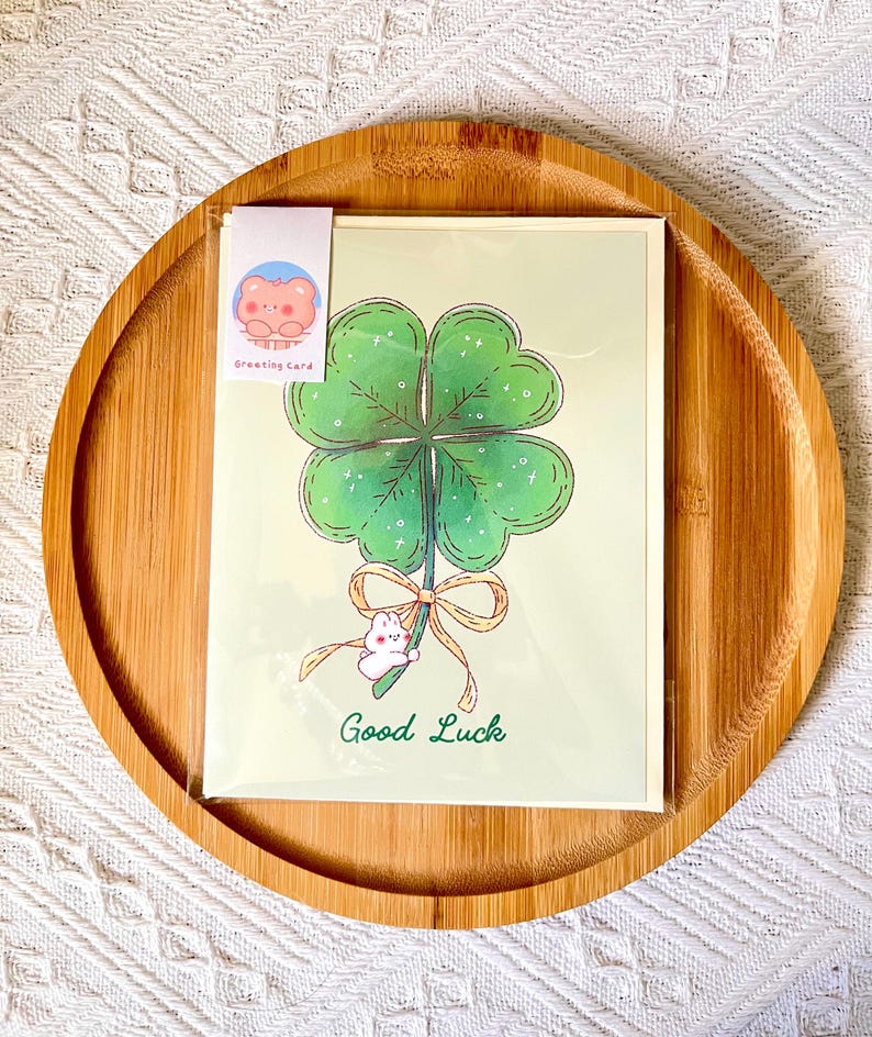 Pu&ograve; includere: Un biglietto di auguri con un quadrifoglio verde, un coniglietto bianco e un fiocco. Le parole "Good Luck" sono scritte in verde. Il biglietto &egrave; su uno sfondo verde chiaro ed &egrave; in una custodia di plastica trasparente. &Egrave; attaccata una piccola etichetta con un orsetto dei cartoni animati.
