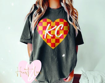Op de afbeelding: Een donkergroen T-shirt met een rood en geel geruit hart met de letters "KC" in wit in het hart.