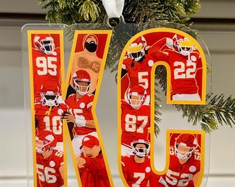 May include: A clear plastic ornament shaped like the letters "KC" with a red background and images of football players wearing red jerseys with white numbers. The ornament has a gold outline.