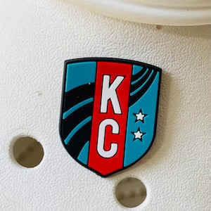 May include: A blue, red, and black shield-shaped charm with the letters "KC" in white and two white stars.
