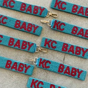 May include: A set of eight beaded keychains with the text "KC BABY" in red beads on a light blue background. The keychains are made of small beads and have a gold metal clasp.