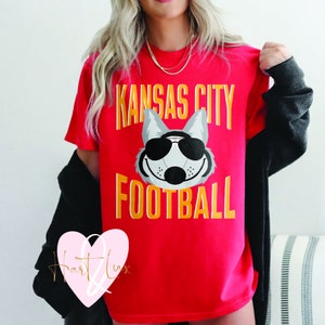 Peut inclure: T-shirt rouge avec un loup gris portant des lunettes de soleil et le texte "Kansas City Football" en jaune.