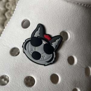 May include: A gray and black cartoon wolf wearing sunglasses and a red hat. The wolf is a shoe charm.