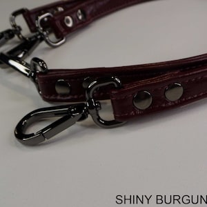 May include: A close-up of a shiny burgundy leather strap with silver metal hardware. The strap has two swivel clasps and three silver rivets. The strap is for a handbag or purse.