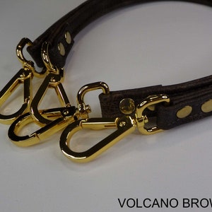 May include: A brown leather strap with gold metal clasps. The strap is designed to be used as a replacement handle for a bag. The strap is labeled "VOLCANO BROWN".