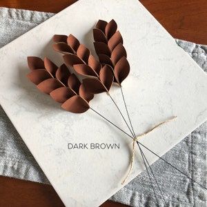 May include: Three dark brown paper wheat stalks tied together with twine. The stalks are made of individual paper leaves folded together. The text "DARK BROWN" is printed on the white surface below the stalks.