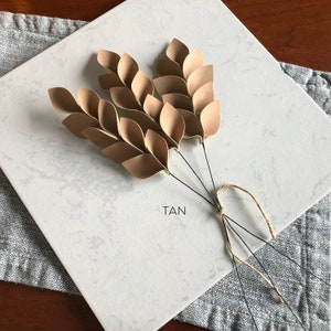 May include: A bouquet of tan paper leaves with wire stems tied together with twine. The leaves are arranged in a fan shape and the stems are trimmed to different lengths. The bouquet is sitting on a white surface with the word "TAN" written on it.