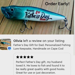 May include: A blue and white fishing lure with a gradient finish, featuring the black text "Happy 1st Father's Day 2026" printed on the side. The lure includes two metal treble hooks attached to the bottom and a realistic eye near the front.