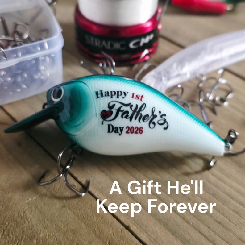May include: A teal and white fishing lure with the text "Happy 1st Father's Day 2026." The lure has a black bill and multiple hooks. The text "A Gift He'll Keep Forever" is also visible. Other fishing accessories are in the background.