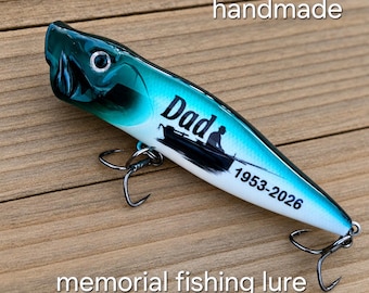 May include: A teal and white fishing lure featuring a black silhouette of a person in a boat, the text "Dad", and the dates "1953-2026". The lure has two metal treble hooks attached to the underside and a single eye on the front.