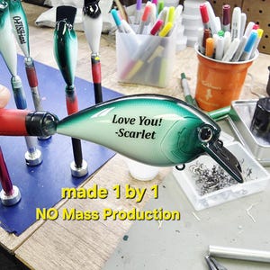 A handmade fishing lure with a personalized message, "Love You!" and "Scarflet", is displayed on a table surrounded by various craft supplies.
