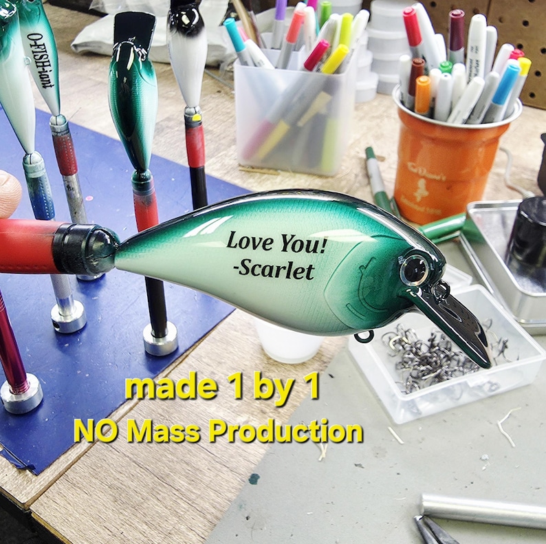 A handmade fishing lure with a personalized message, "Love You!" and "Scarflet", is displayed on a table surrounded by various craft supplies.