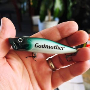 May include: A fishing lure with a gradient finish transitioning from dark green at the head to a pale white body. The word Godmother is printed in black text on the side. The lure features a black head with a white eye and two metal treble hooks.