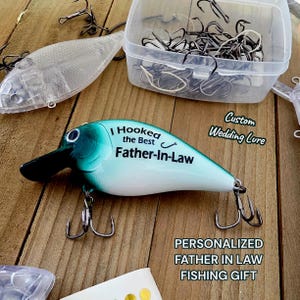 a fishing lure with a personalized message, a clear plastic container filled with fishing hooks, and a wooden surface with a sign that reads "Custom Wedding Lure".