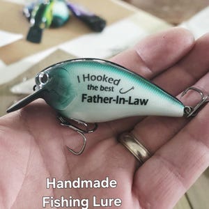 A hand holding a handmade fishing lure with the text "I Hooked the best Father-in-Law" printed on it.