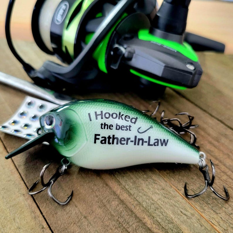 A fishing lure with the text "I Hooked the best Father-in-Law" on it, along with a fishing rod and other fishing gear, is displayed on a wooden surface.
