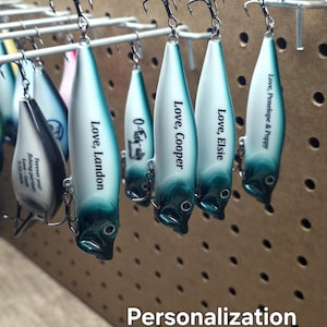May include: A collection of fishing lures with a white and teal gradient finish, each featuring a metal hook and personalized text including names like Landon, Cooper, Elsie, and Penelope & Poppy, with the text Personalization Included on side #2.