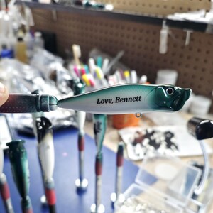 May include: A fishing lure with a white and dark green gradient body featuring the black text Love, Bennett printed on the side. The lure has a pointed nose, a small eye detail, and a metal attachment loop on the underside of the main body.