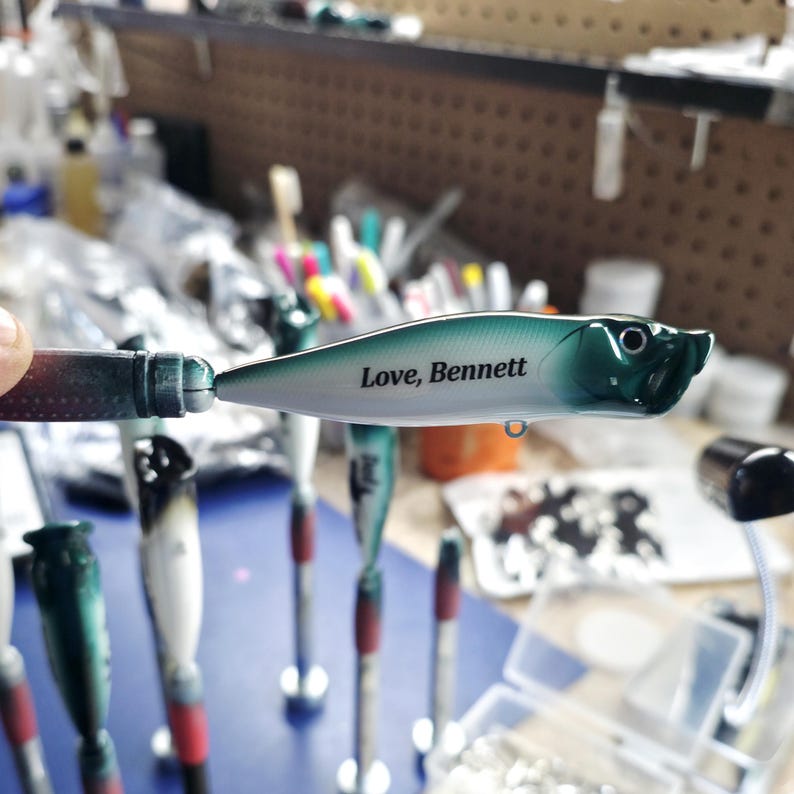 May include: A fishing lure with a white and dark green gradient body featuring the black text Love, Bennett printed on the side. The lure has a pointed nose, a small eye detail, and a metal attachment loop on the underside of the main body.