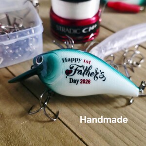 May include: A handmade fishing lure with a white and teal body, featuring the text "Happy 1st Father's Day 2026." The lure has a dark green bill and two treble hooks. Other fishing supplies are in the background.
