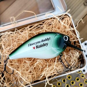 May include: A fishing lure with a green and white body, black beak, and two treble hooks. The lure is in a metal tin filled with straw. Text on the lure reads "I love you daddy!" and "Kaisley" with a heart symbol.