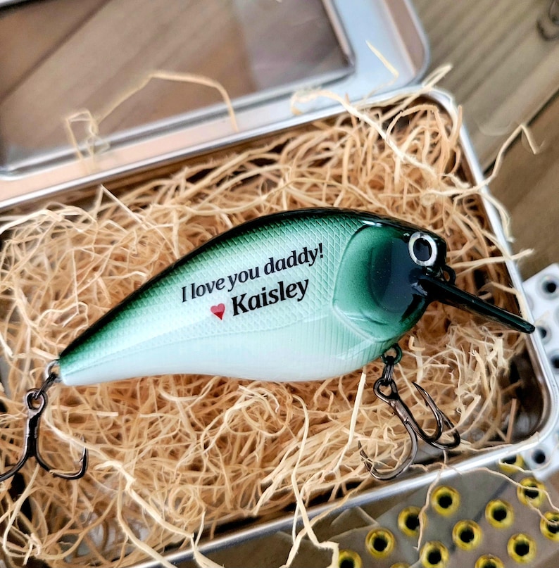 May include: A fishing lure with a green and white body, black beak, and two treble hooks. The lure is in a metal tin filled with straw. Text on the lure reads "I love you daddy!" and "Kaisley" with a heart symbol.