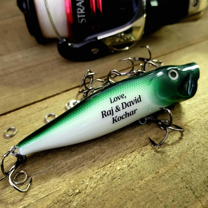 May include: A green and white fishing lure featuring the printed text Love, Raj & David Kochar. The lure has a pointed head, a smooth body, and multiple metal treble hooks attached to the underside, positioned near a black and red fishing reel.