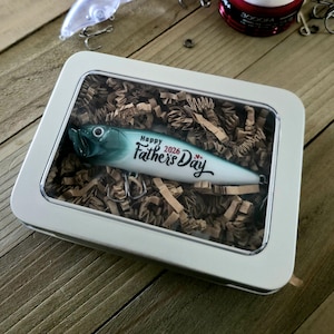 May include: A white rectangular metal tin with a clear window lid containing a fishing lure nestled in brown paper filler. The lure is white and teal with the black text Happy Father's Day 2026 printed on the side and silver metal hooks.