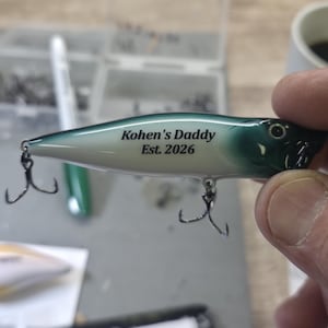May include: A fishing lure with a gradient green and white body featuring the black printed text Kohen's Daddy Est. 2026. The lure includes two silver metal treble hooks attached to the underside and a small eye detail on the front section.