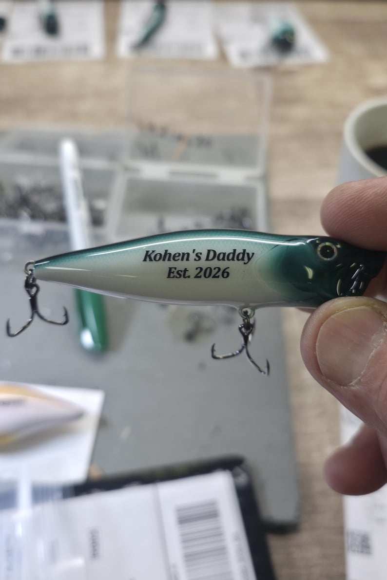 May include: A fishing lure with a gradient green and white body featuring the black printed text Kohen's Daddy Est. 2026. The lure includes two silver metal treble hooks attached to the underside and a small eye detail on the front section.