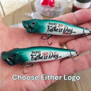 May include: Two fishing lures with a green and white gradient finish. The top lure features the text "Happy 1st Father's Day 2026" in black script. The bottom lure displays "Happy Father's Day 2026" with small red hearts. Both lures have metal hooks.