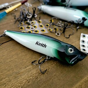 May include: A fishing lure with a white and dark green gradient body featuring the name Keith printed in black. The lure has a black head, a single eye, and two metal treble hooks attached to the underside. Additional hooks and eyes are visible.