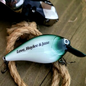 May include: A fishing lure with a white and green body, black beak, and a silver hook. The text "Love, Haylee & Jase" is printed on the side. A fishing reel and rope are in the background.