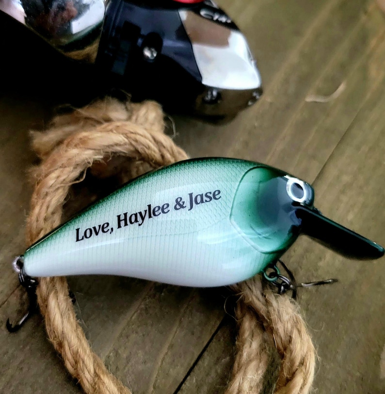May include: A fishing lure with a white and green body, black beak, and a silver hook. The text "Love, Haylee & Jase" is printed on the side. A fishing reel and rope are in the background.