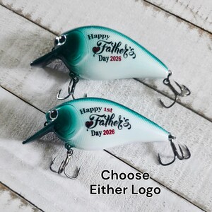 May include: Two fishing lures in teal and white. Each lure features the text "Happy Father's Day 2026". One lure says "Happy 1st Father's Day 2026". The text "Choose Either Logo" is also present. The lures have hooks and a beak-like projection.