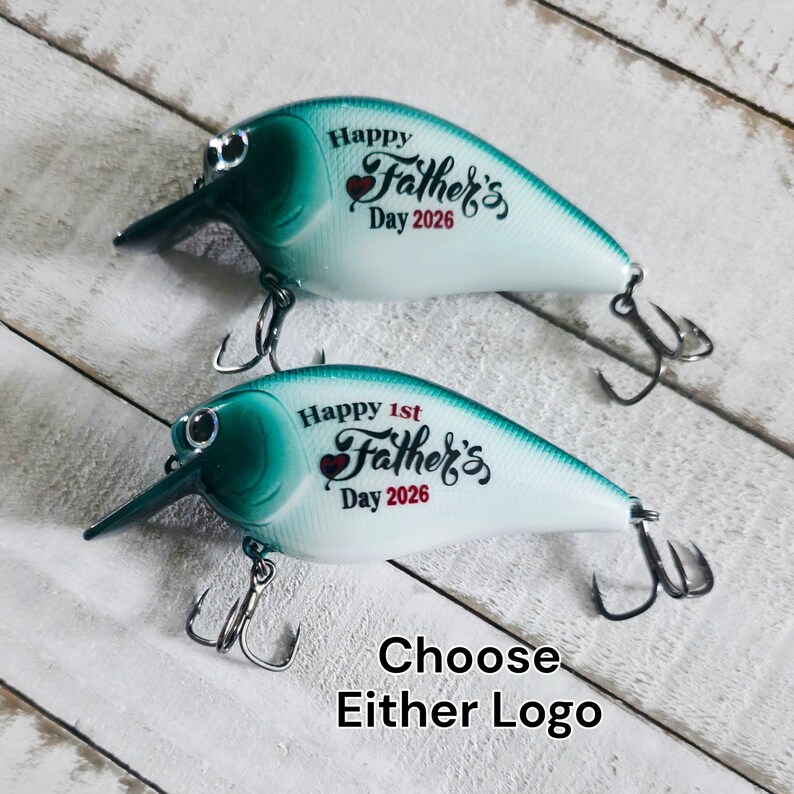 May include: Two fishing lures in teal and white. Each lure features the text "Happy Father's Day 2026". One lure says "Happy 1st Father's Day 2026". The text "Choose Either Logo" is also present. The lures have hooks and a beak-like projection.