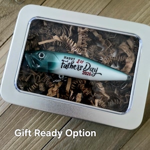 May include: A fishing lure with a green and white gradient finish, featuring the black text Happy 1st Father's Day 2026, encased in a rectangular silver metal tin with a clear lid, resting on a bed of brown shredded paper filler material.