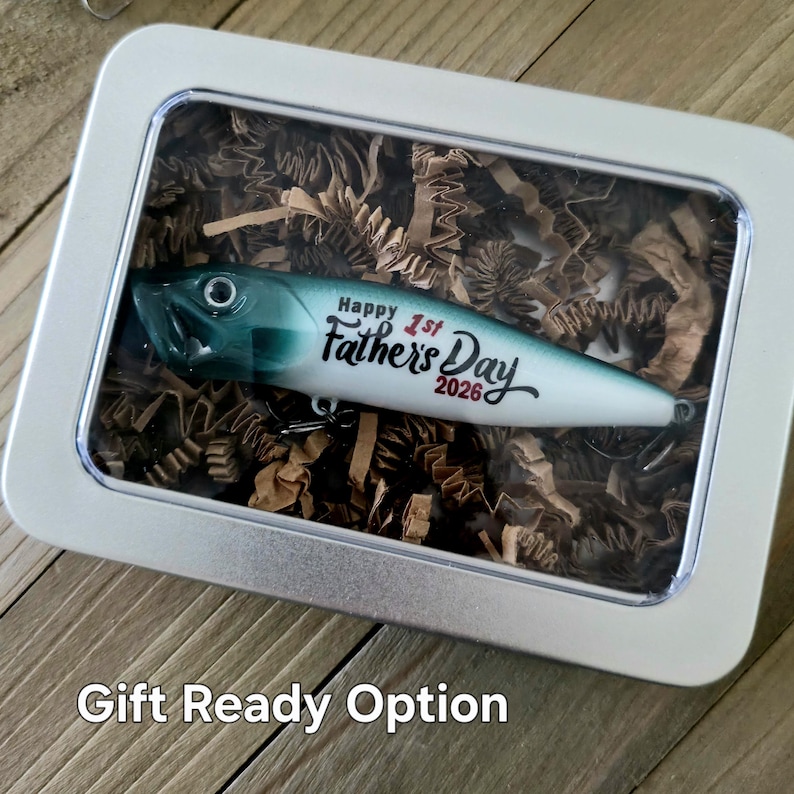 May include: A fishing lure with a green and white gradient finish, featuring the black text Happy 1st Father's Day 2026, encased in a rectangular silver metal tin with a clear lid, resting on a bed of brown shredded paper filler material.