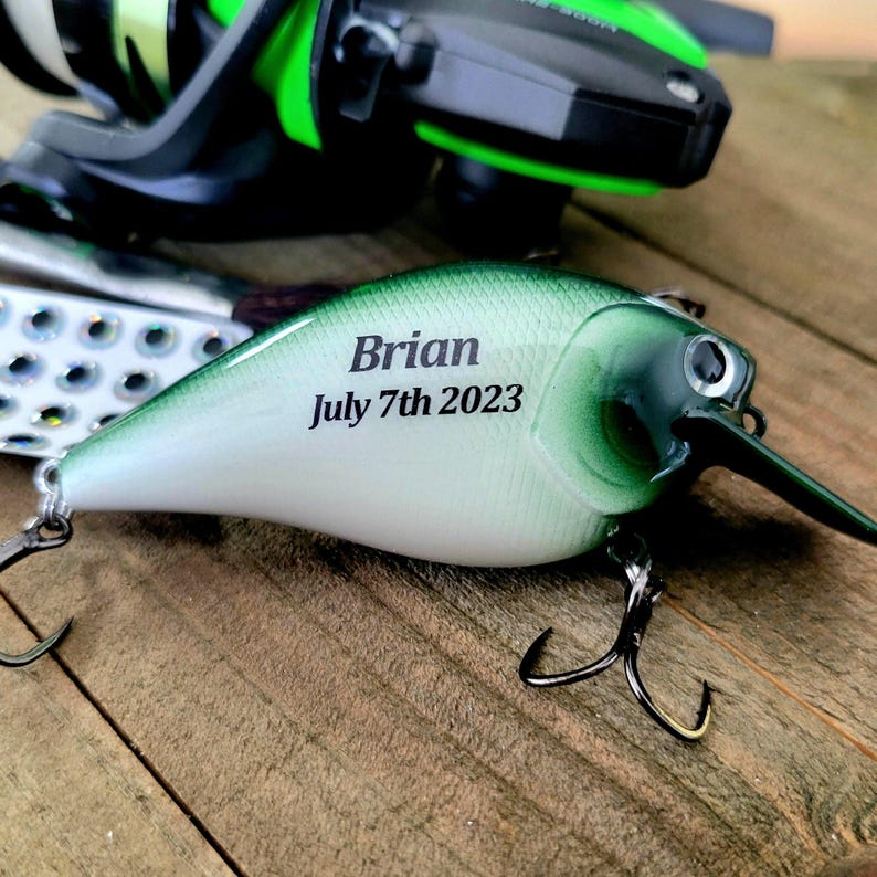 A personalized fishing lure with the name "Brian" and the date "July 7th 2023" engraved on it, resting on a wooden surface.