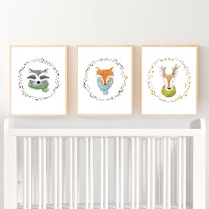 May include: Three framed watercolor illustrations of woodland animals wearing scarves. The illustrations feature a raccoon, a fox, and a deer, each surrounded by a wreath of leaves and flowers.
