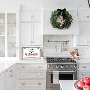 May include: A white kitchen featuring a Christmas wreath and a sign reading "North Pole Milk & Cookie Co. Baking Santa's Favourites". The wreath is green with a dark green bow and gold bells. The kitchen has white cabinets and a stainless steel cooker.