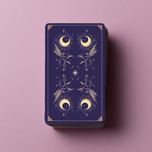 Puede incluir: Una baraja de cartas de tarot rectangular con un dise&ntilde;o de reverso en azul marino profundo, con ilustraciones en color dorado de cuatro lunas crecientes, alas estilizadas, enredaderas florales y peque&ntilde;as estrellas en un patr&oacute;n sim&eacute;trico.