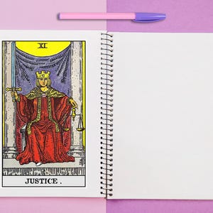 Puede incluir: Un cuaderno de espiral con una ilustraci&oacute;n de la carta del tarot Justicia en la p&aacute;gina izquierda, que muestra una figura con t&uacute;nica roja sosteniendo una espada y una balanza. Un bol&iacute;grafo rosa claro descansa horizontalmente sobre las p&aacute;ginas.