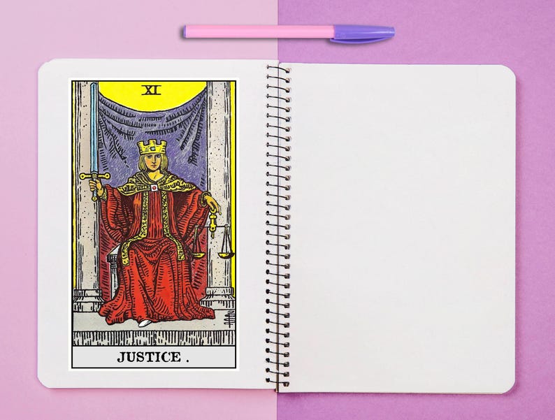 Puede incluir: Un cuaderno de espiral con una ilustraci&oacute;n de la carta del tarot Justicia en la p&aacute;gina izquierda, que muestra una figura con t&uacute;nica roja sosteniendo una espada y una balanza. Un bol&iacute;grafo rosa claro descansa horizontalmente sobre las p&aacute;ginas.