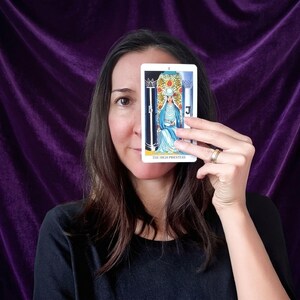 May include: A rectangular tarot card titled The High Priestess featuring a central figure in blue robes seated between two pillars, one black and one white, with a crown and a pomegranate patterned backdrop, held up against a dark purple fabric background.