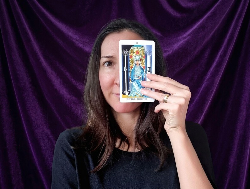 May include: A rectangular tarot card titled The High Priestess featuring a central figure in blue robes seated between two pillars, one black and one white, with a crown and a pomegranate patterned backdrop, held up against a dark purple fabric background.