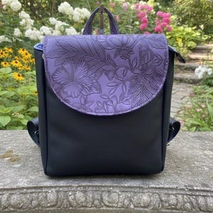 May include: A black backpack with a purple flap featuring a floral design. The backpack has a top handle and adjustable straps. The background includes a variety of colorful flowers.