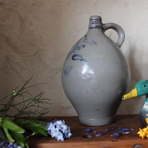 Puede incluir: Una jarra de cerámica gris con diseños pintados en azul y blanco se encuentra sobre una superficie de madera junto a un pato de madera pintado de verde y azul. El pato tiene un pico amarillo y mira hacia el lado izquierdo de la imagen.