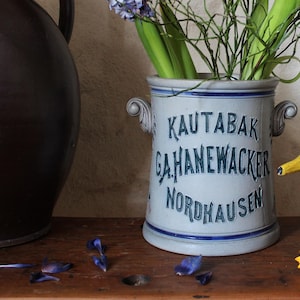 以下が含まれることがあります： 前面に「Kauttabak Gahnewacker Nordhausen」と書かれた青と白の陶器の壺。壺には紫色のヒヤシンスと緑色の茎が詰められています。緑と黒の塗料で塗られた木製の鴨が壺の隣の木の表面に座っています。