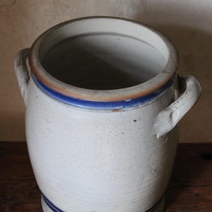 Può includere: Un vaso di ceramica bianca con due manici e un bordo blu e marrone. Il vaso è vuoto e si trova su una superficie di legno.